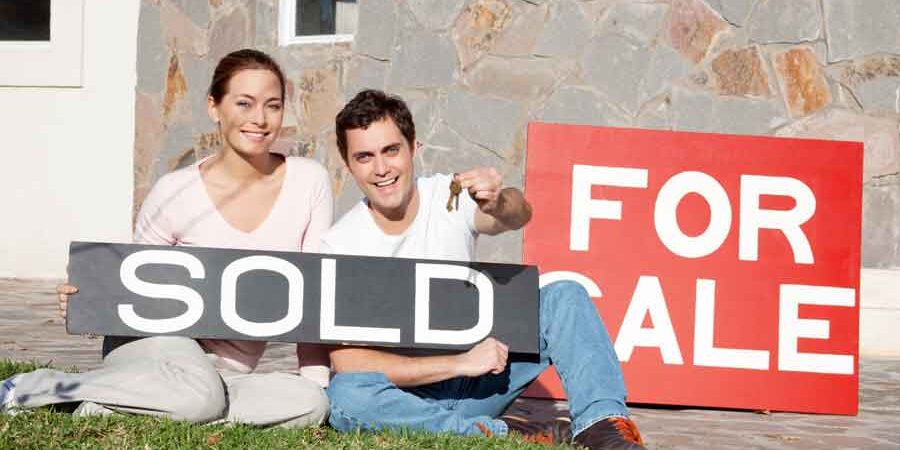 seller’s-market young couple in front of a house with a for sale sign and holding a sold sign. It's a seller's market